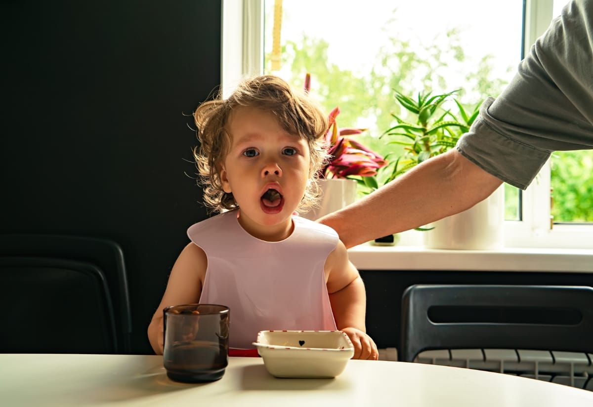 Young child showing signs of choking while seated at a dining table, with an adult’s arm reaching towards them