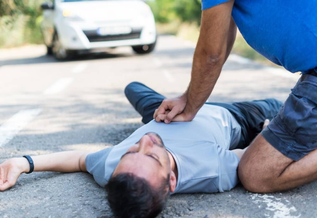 Eine Person, die eine Herz-Lungen-Wiederbelebung an einem bewusstlosen Mann durchführt, der in der Nähe eines Autos auf der Straße liegt, veranschaulicht die Herz-Lungen-Wiederbelebungsausbildung im Zusammenhang mit der theoretischen Fahrprüfung.